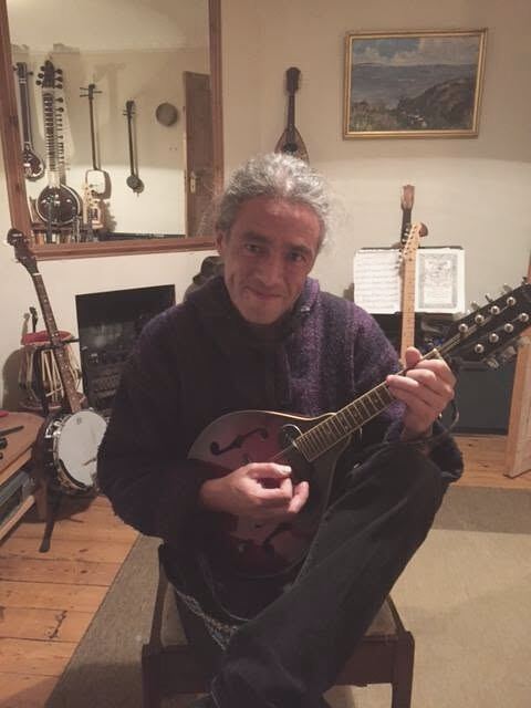 Tom Ashton seated with a mandolin, smiling at the camera, in a room hung with stringed instruments — a sitar among them, reflected in a mirror — with a music stand and a framed landscape painting behind him.