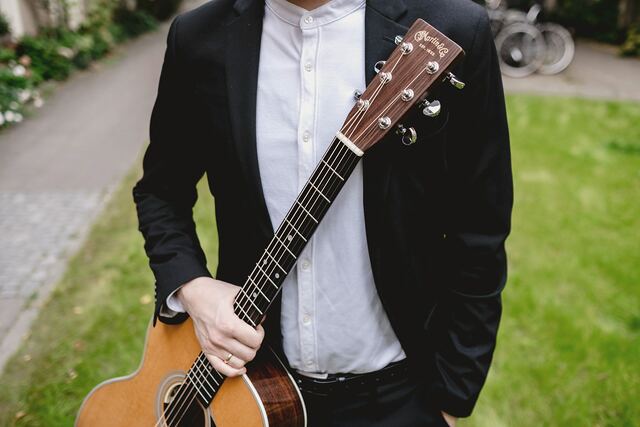 A man in a dark suit and white shirt holding an acoustic guitar by its neck, outdoors on a lawn, cropped below the face.