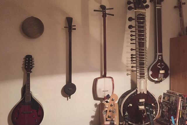 Stringed instruments hanging on a plain wall: a red sunburst mandolin, two long-necked lutes with skin-covered bodies, a sitar, and beside it a smaller instrument with an inlaid teardrop body; the headstock of a bass guitar and a row of books below.